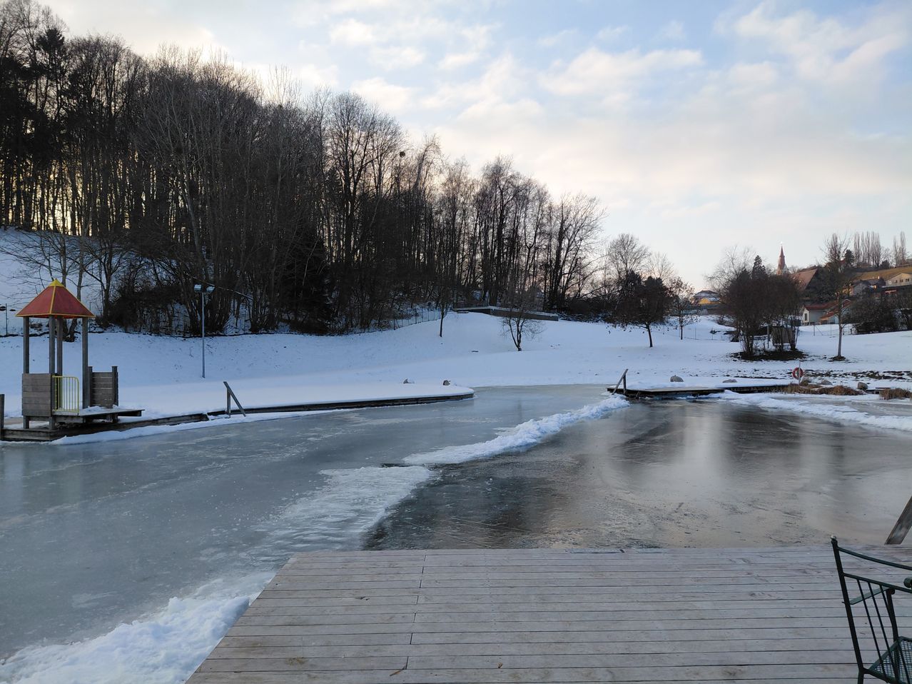 Ein gefrorener See mit einem Holzsteg, umgeben von schneebedeckten Bäumen und Hügeln. Ein Gebäude ist in der Ferne sichtbar.