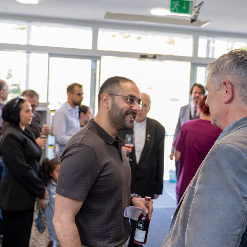 Eine Gruppe von Menschen bei einer gesellschaftlichen Veranstaltung. Ein Mann mit Brille hält eine Flasche und unterhält sich mit einem anderen Mann. Eine Frau mit Handtasche steht in der Nähe. Andere Teilnehmer unterhalten sich im Hintergrund.