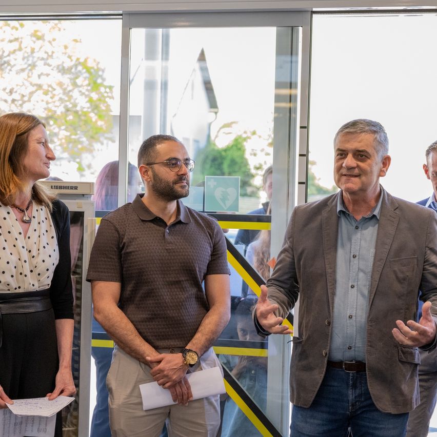 Eine Gruppe von Menschen steht vor einer Glastür, zwei Männer in Freizeitkleidung, einer in einem Anzug und eine Frau mit einer Halskette. Sie lächeln und scheinen ein Gespräch zu führen.