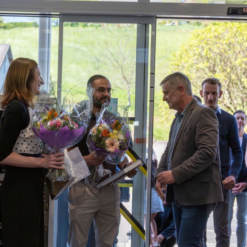 Eine Frau mit einem Blumenstrauß steht neben einem Mann, der ein Buch und einen weiteren Strauß hält. Sie stehen vor einer Glastür. Hinter ihnen schaut ein Mann sie an.