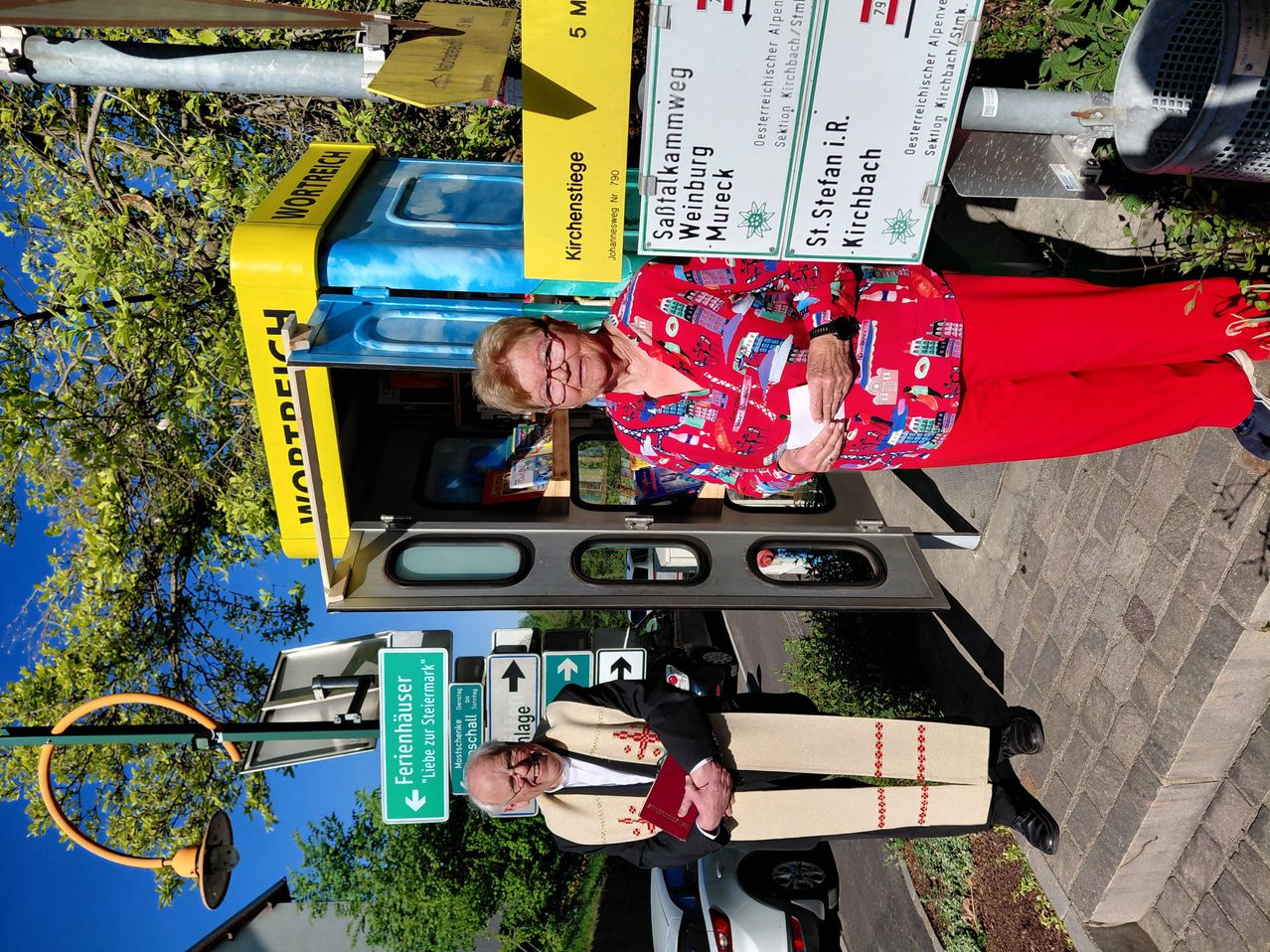 Zwei ältere Personen stehen vor einem gelben Kiosk mit einem Schild, das Wortreich anzeigt. Eine Frau hält ein Papier, während ein Mann ein Buch hält. Dahinter befindet sich ein Straßenschild, das 5 M Entfernung anzeigt.
