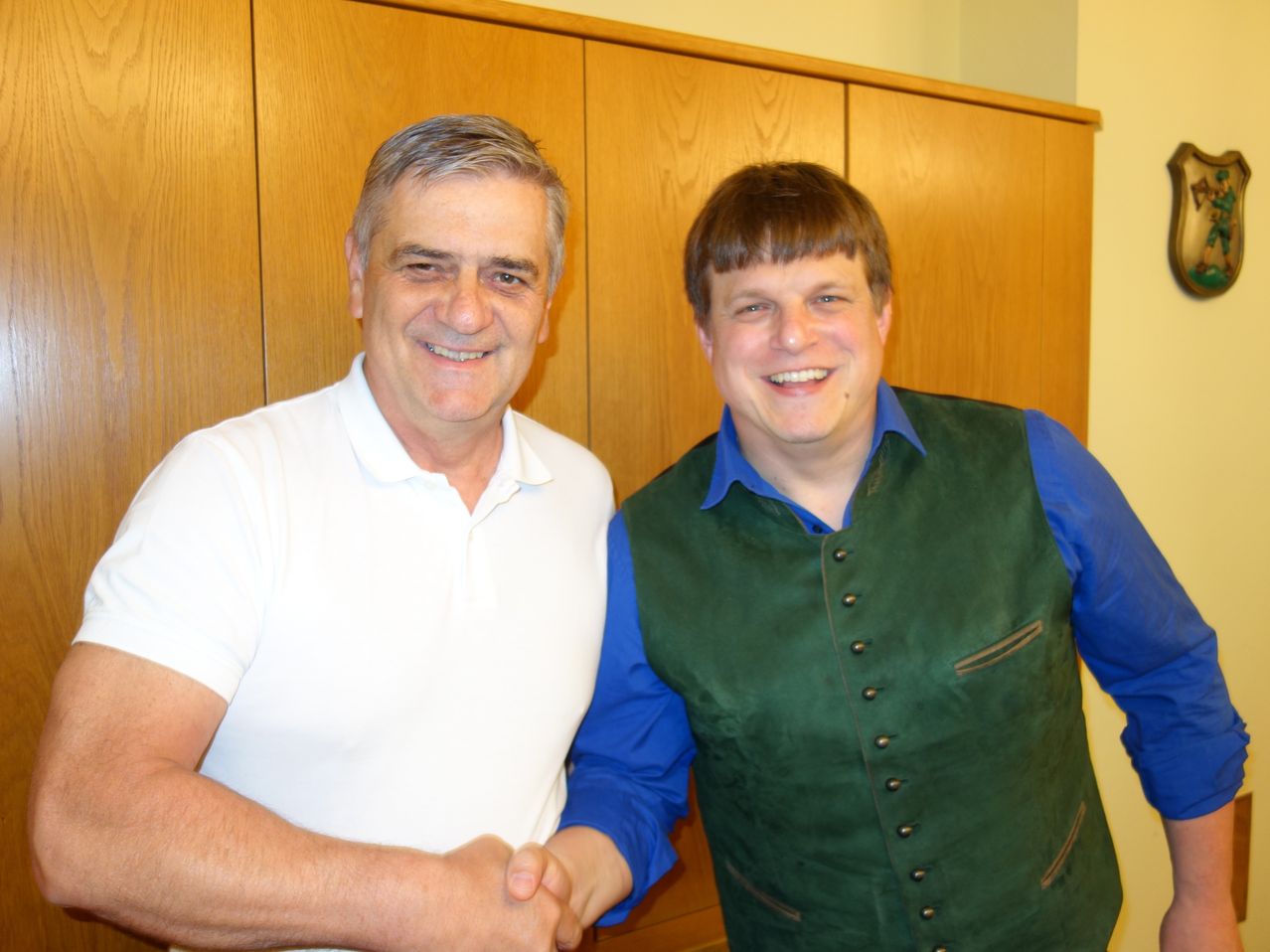 Zwei Männer stehen nebeneinander und lächeln. Der Mann auf der linken Seite trägt ein weißes Poloshirt. Der Mann auf der rechten Seite trägt eine grüne Weste über einem blauen Hemd. Sie stehen vor einem Holzschrank.