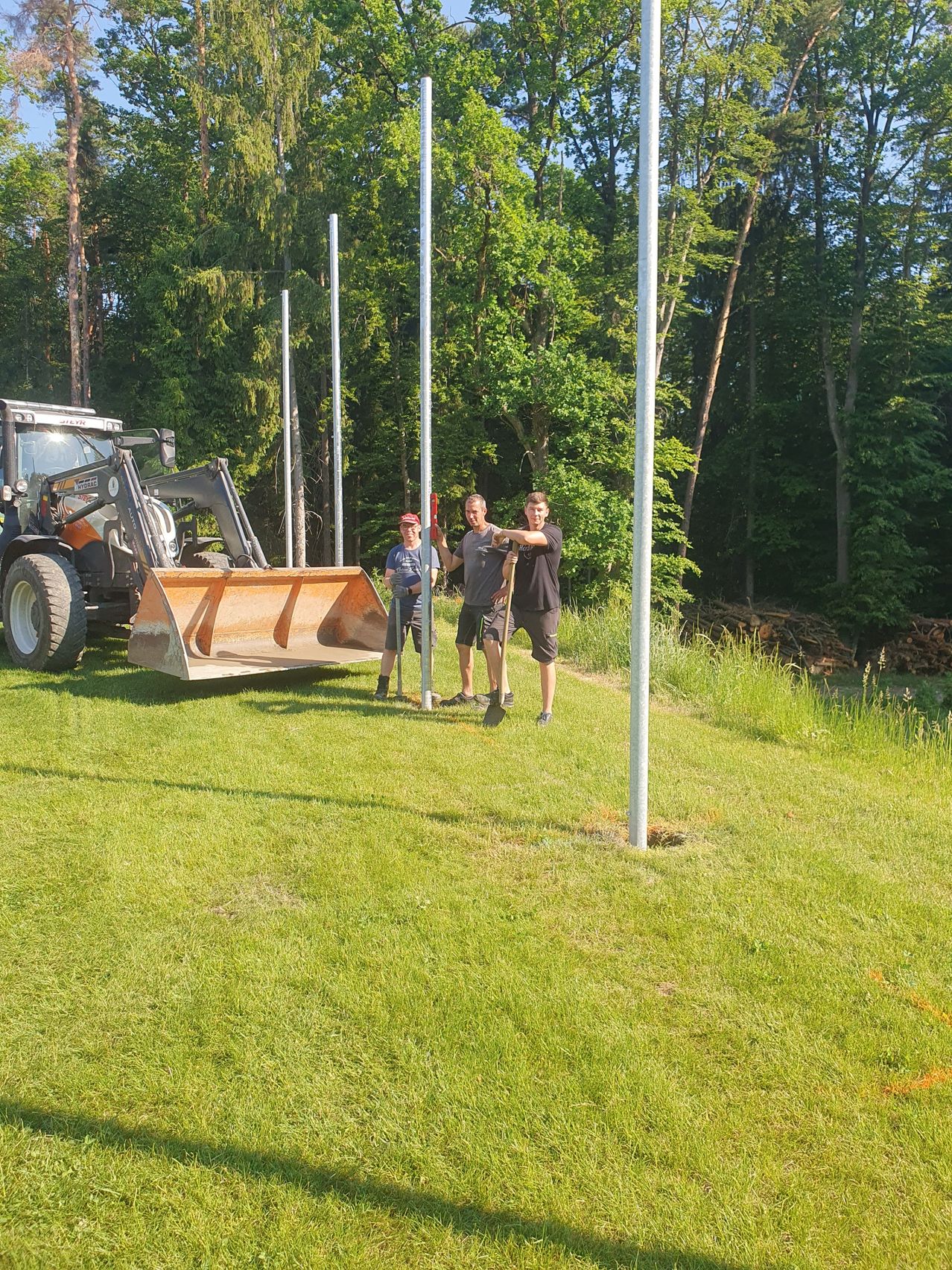 Drei Männer richten Stangen auf einer Rasenfläche auf, mit einem Traktor im Hintergrund. Sie sind von einem dichten Wald umgeben.