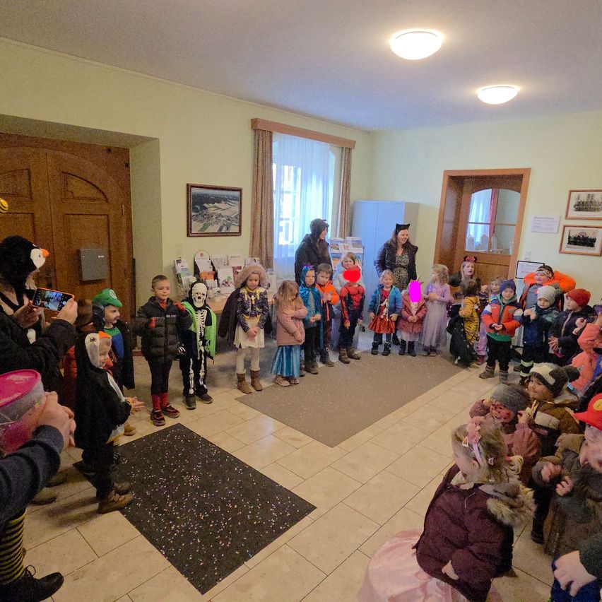 Eine Gruppe von Kindern in verschiedenen Kostümen versammelt sich in einem Raum, einige halten Requisiten. Zwei Erwachsene stehen unter ihnen, einer trägt einen Hut. Der Raum hat weiße Wände, geflieste Böden und zwei Türen. Es gibt Bilderrahmen und eine Lampe an der Wand.
