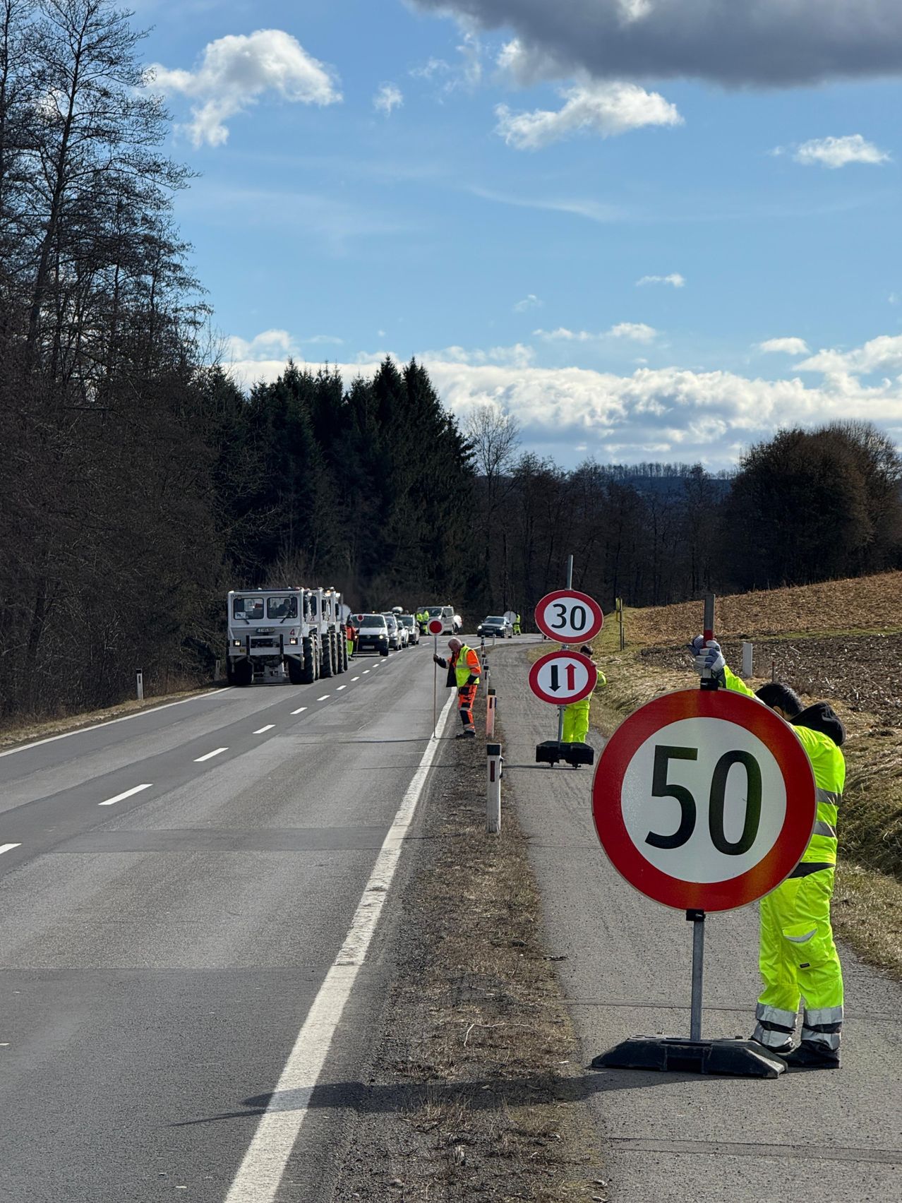 Eine Straßenbaumannschaft stellt Geschwindigkeitsbegrenzungsschilder auf einer zweispurigen Straße ein, an der Fahrzeuge vorbeifahren. Im Hintergrund befinden sich Bäume und ein bewölkter Himmel.