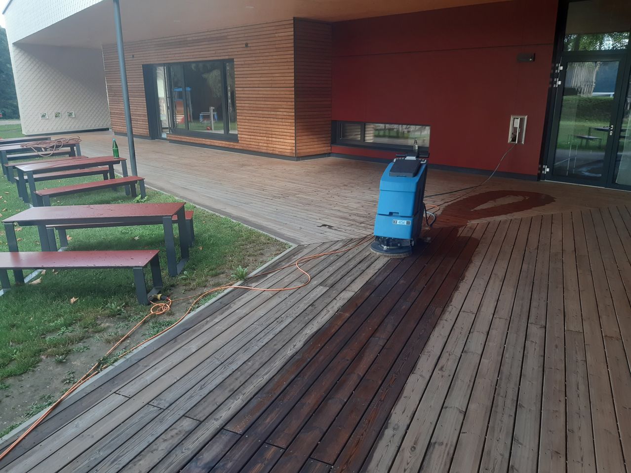 Eine blaue Reinigungsmaschine wird verwendet, um eine Holzterrasse zu reinigen. Die Terrasse ist von einer roten Wand umgeben und es gibt Bänke und einen Pfosten in der Nähe.