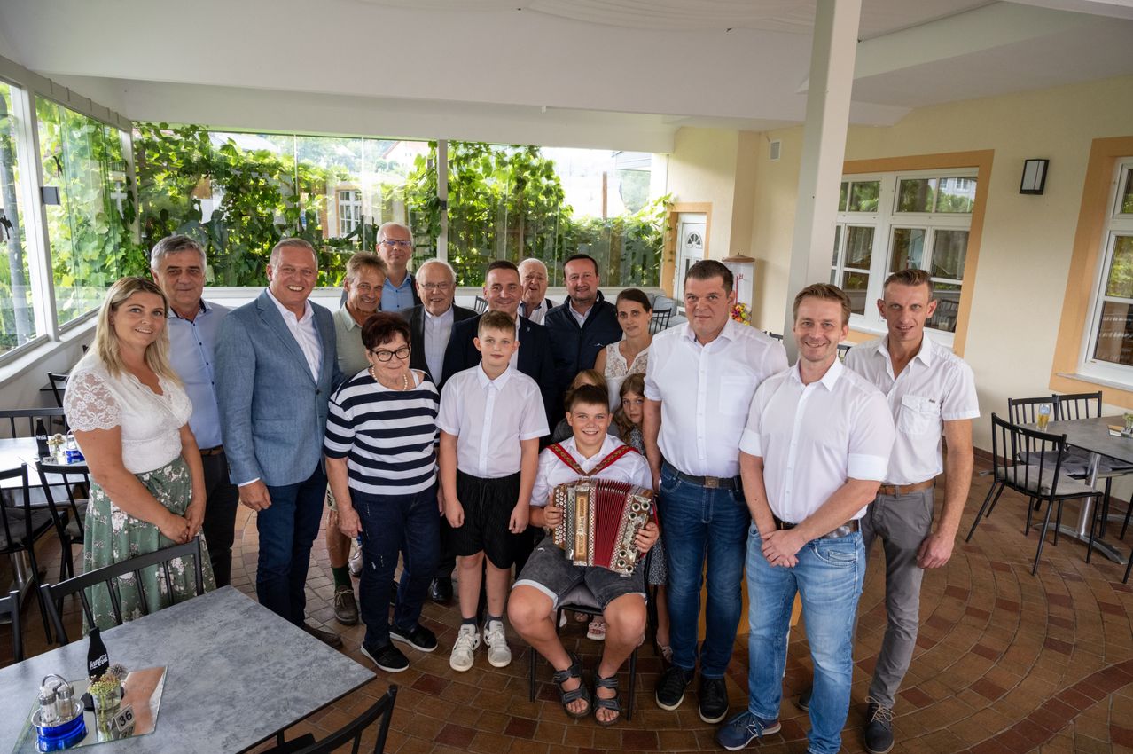 Eine Gruppe von Menschen, einschließlich Erwachsener und Kindern, posiert für ein Foto in einem Raum mit gefliestem Boden und Glaswänden. Einige lächeln, und ein Junge sitzt auf einem Stuhl und spielt Akkordeon.