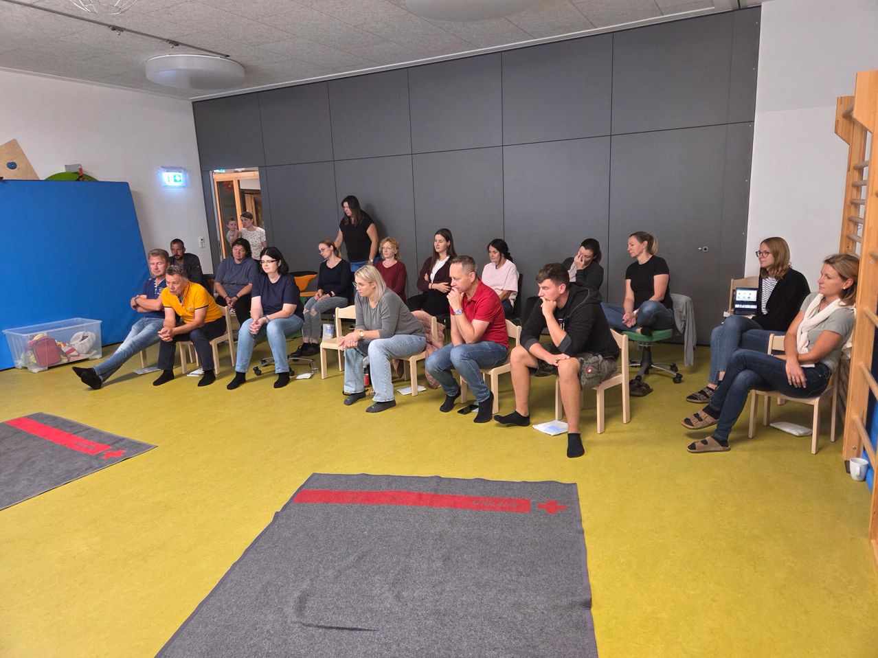 Eine Gruppe von Menschen sitzt in einem Kreis auf Stühlen in einem Raum, einige tragen Brillen und Turnschuhe, mit einem Teppich auf dem Boden.