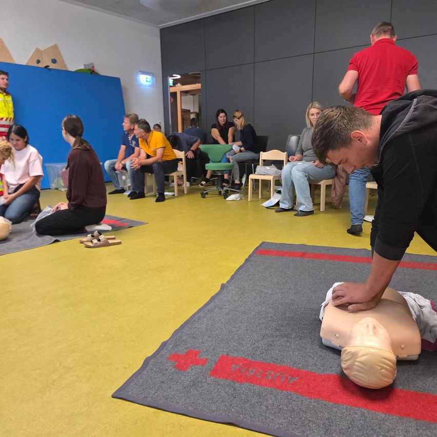 Eine Gruppe von Menschen ist in einem Raum für eine CPR-Schulung versammelt. Ein Mann demonstriert CPR an einem Dummy.