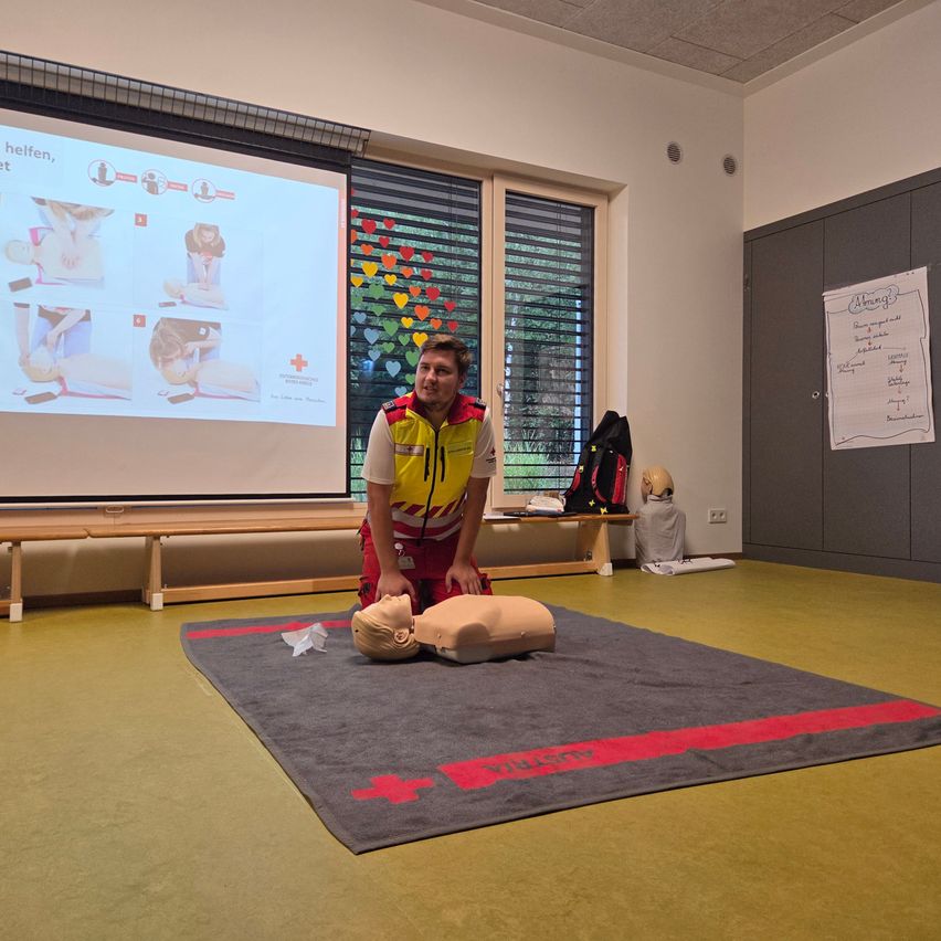 Ein Mann in einem gelben und roten Sicherheitsweste demonstriert CPR an einem Mannequin in einem Klassenzimmer. Eine Projektionsleinwand hinter ihm zeigt CPR-Anweisungen.