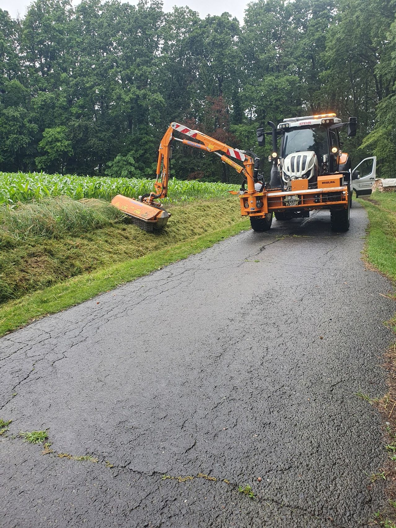 Ein orangefarbener Traktor mit einer Grasschneidemaschine steht auf einer Straße. Im Hintergrund sind Bäume und Maisfelder zu sehen.