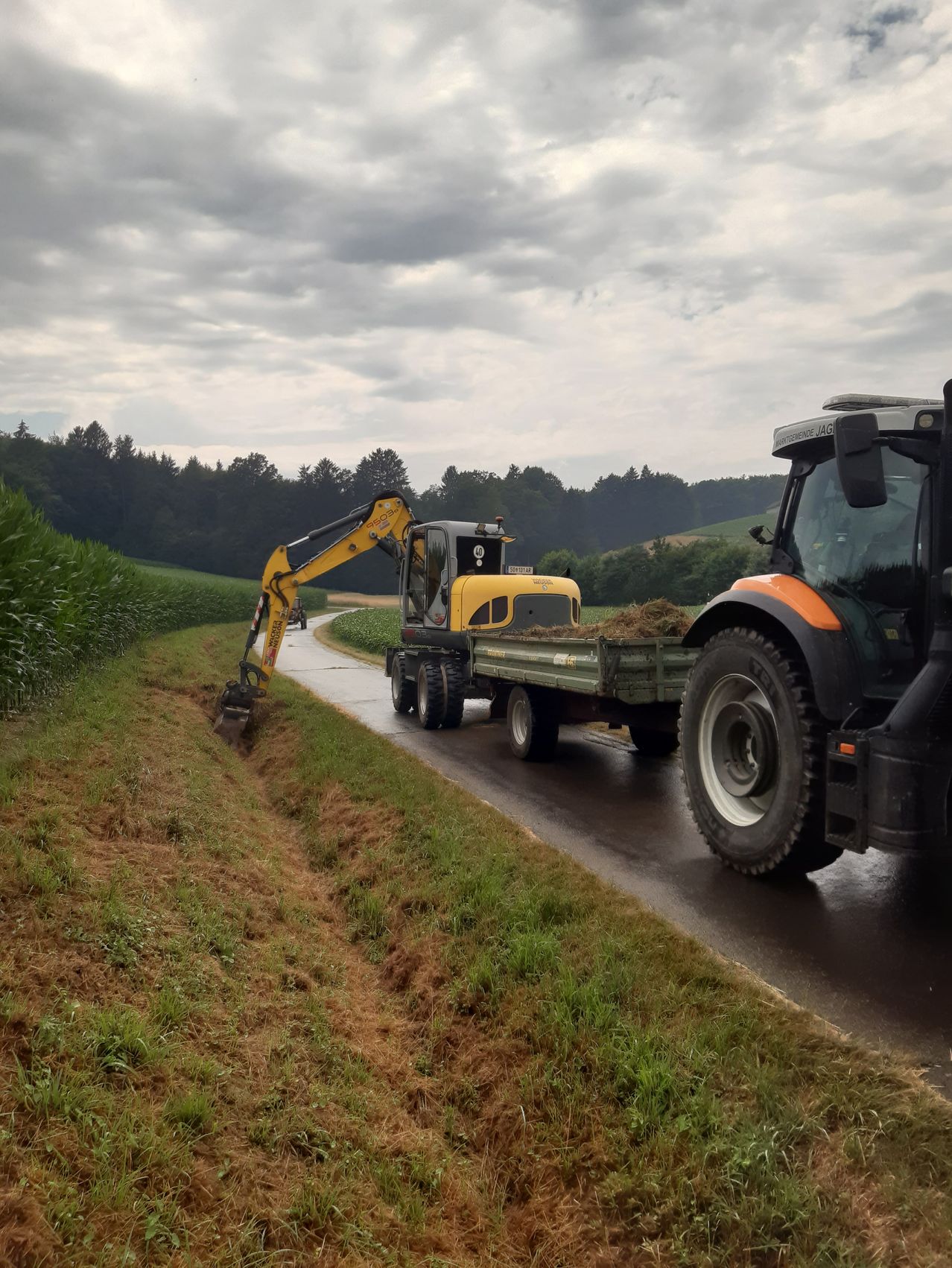 A yellow excavator is on the road, loading hay into a trailer. A tractor follows. The road is lined with grass and trees.