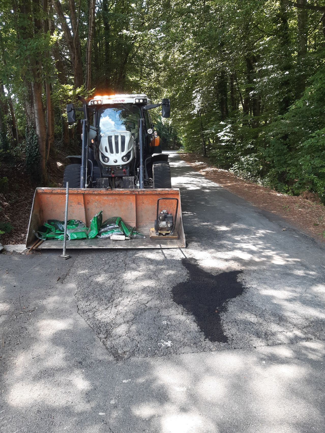 A tractor with a snowplow is parked on the side of the road. It has several bags of cement on its front. The road is surrounded by trees and appears to be under construction.