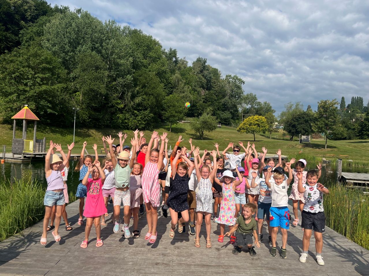 A group of children and adults are jumping and waving on a wooden dock by a lake, with trees and a cloudy sky in the background.