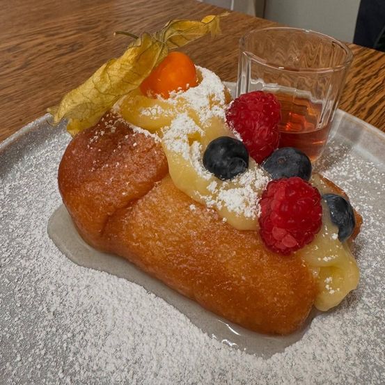 Ein Gebäck mit goldbrauner Kruste, belegt mit verschiedenen Früchten und einer Prise Puderzucker, begleitet von einem kleinen Glas Sirup auf einem Holztisch.