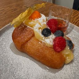 Ein Gebäck mit goldbrauner Kruste, belegt mit verschiedenen Früchten und einer Prise Puderzucker, begleitet von einem kleinen Glas Sirup auf einem Holztisch.