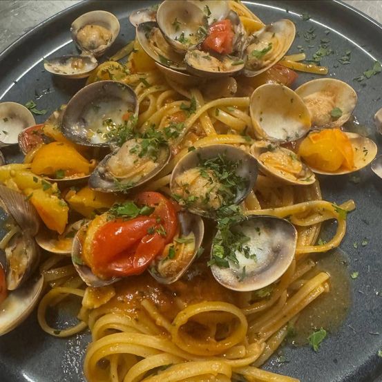 Ein Teller mit Pasta, Muscheln, Kirschtomaten und Kräutern. Die Pasta ist orange und die Muscheln sind geöffnet.