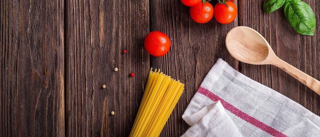 Auf einem Holztisch befinden sich ein Holzlöffel, ein Basilikumbüschel, eine Traube Kirschtomaten und ein Spaghetti-Bündel. Ein weißes Handtuch mit rotem Streifen ist ebenfalls auf dem Tisch.
