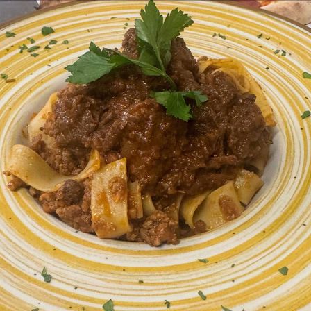 Ein Teller mit Rinderragout und Pappardelle-Pasta, garniert mit frischem Petersilie. Das Gericht wird auf einem weißen Teller mit gelben Streifen serviert.