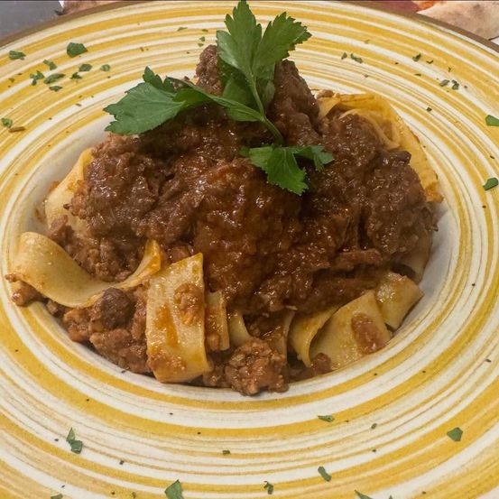 Ein Teller mit Rinderragout und Pappardelle-Pasta, garniert mit frischem Petersilie. Das Gericht wird auf einem weißen Teller mit gelben Streifen serviert.