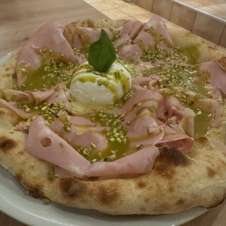 Eine Pizza mit Schinkenscheiben, einer Mozzarellakugel, Pistazien und einem Basilikumblatt auf einem weißen Teller.