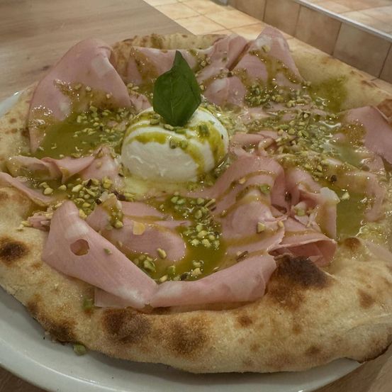 Eine Pizza mit Schinkenscheiben, einer Mozzarellakugel, Pistazien und einem Basilikumblatt auf einem weißen Teller.