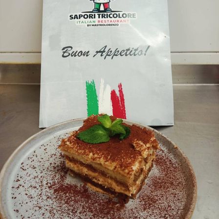 Ein Stück Tiramisu auf einem Teller mit einer Minzblatt, bestreut mit Kakaopulver, mit einem Restaurantbeutel im Hintergrund.