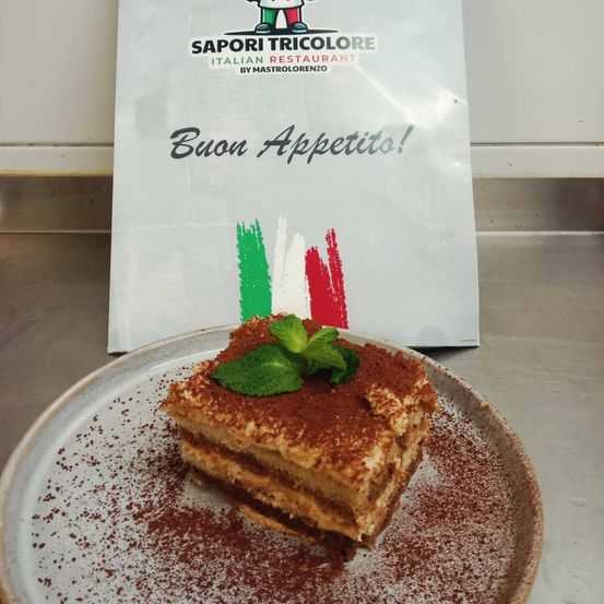 Ein Stück Tiramisu auf einem Teller mit einer Minzblatt, bestreut mit Kakaopulver, mit einem Restaurantbeutel im Hintergrund.