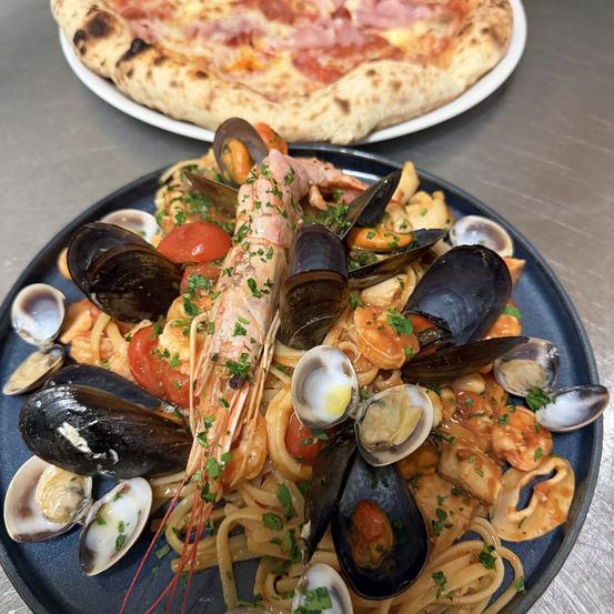 Ein Meeresfrüchte-Pastagericht mit Muscheln, Garnelen und Tomaten auf einem blauen Teller, mit einer Pizza im Hintergrund.
