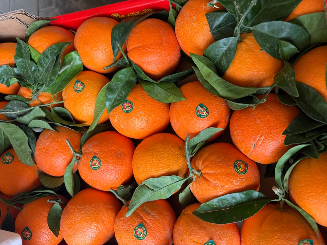 Eine Nahaufnahme von einer Ansammlung von Orangen mit grünen Blättern, in einem roten Behälter platziert. Jede Orange hat einen grünen Aufkleber.