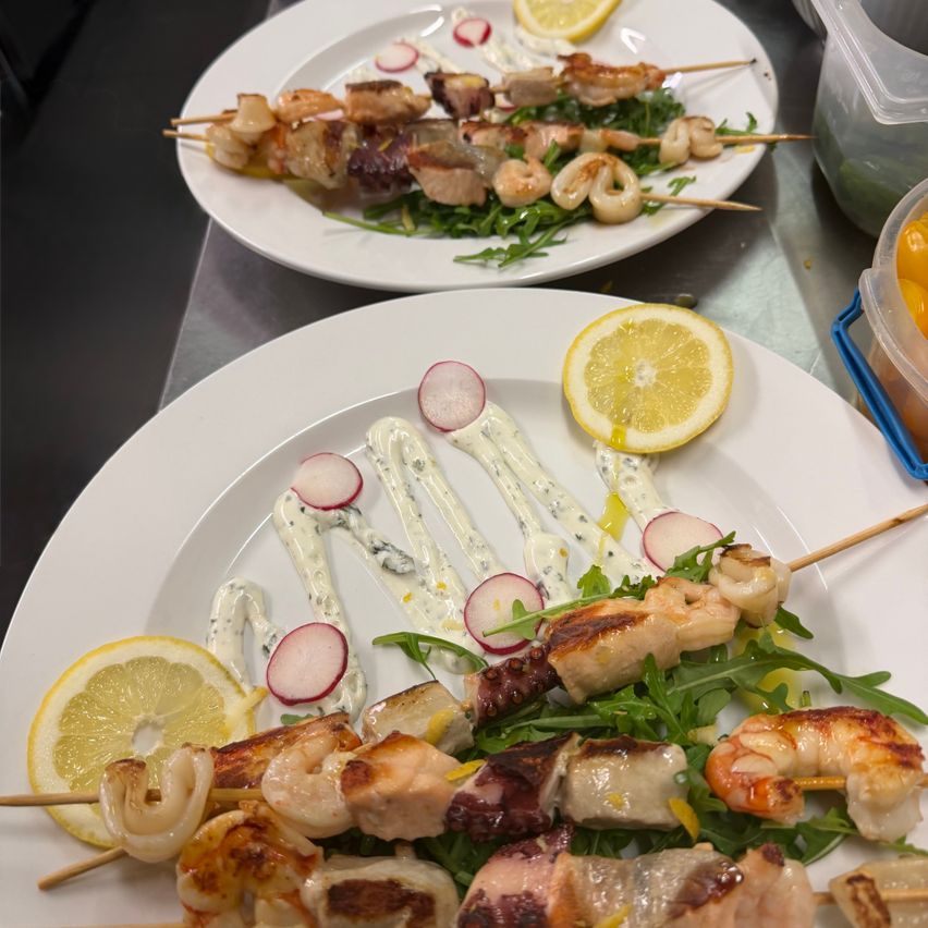 Zwei Teller mit Spießen aus Meeresfrüchten und Gemüse, serviert mit Rucola, Zitronenscheiben und Blauschimmelkäse-Sauce. Behälter und Schüsseln im Hintergrund.