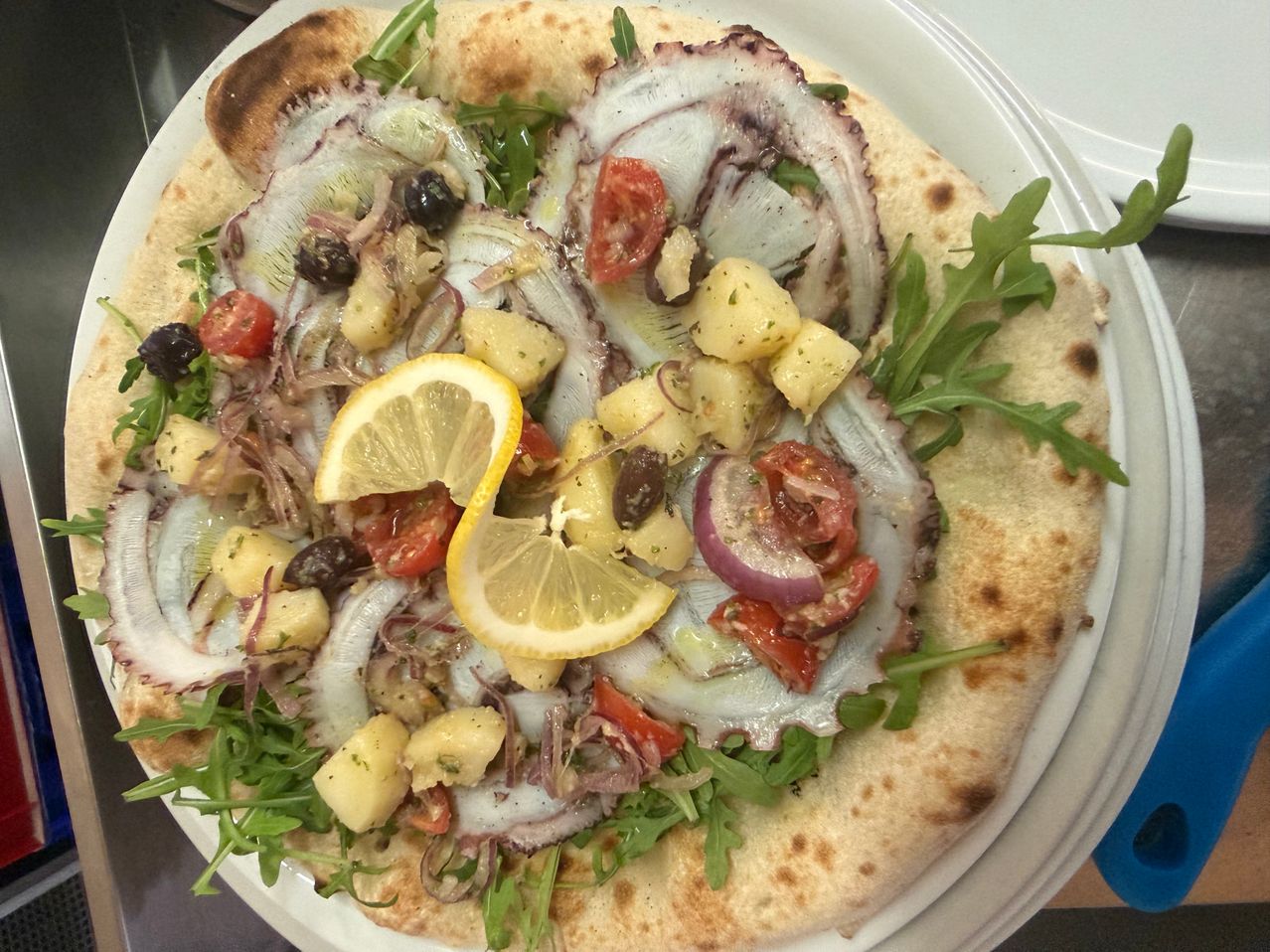 Eine Flatbread-Pizza mit einer Zitronenscheibe, Oktopus, Kartoffeln, Tomaten und Rucola auf einem weißen Teller.