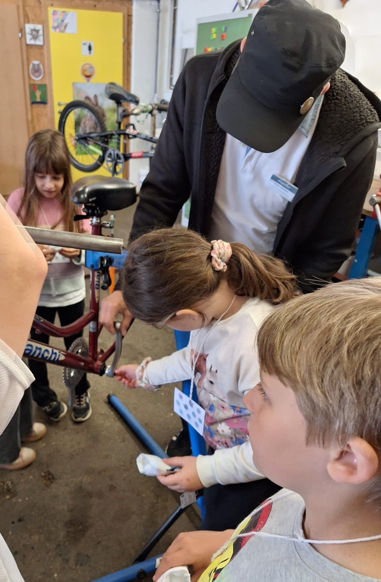 Mehrere Kinder und ein Erwachsener arbeiten an einem Fahrrad. Ein Kind justiert den Sattel, während ein anderes Kind das Lenker hält. Ein Mann in einer Kappe hilft ihnen.