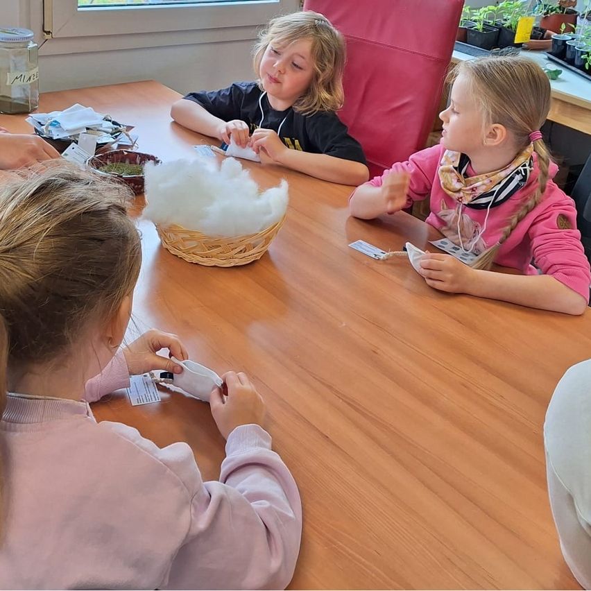 Vier junge Mädchen sitzen um einen Tisch herum, auf dem ein Korb mit Baumwolle steht. Sie scheinen an einer Bastelaktivität beteiligt zu sein.