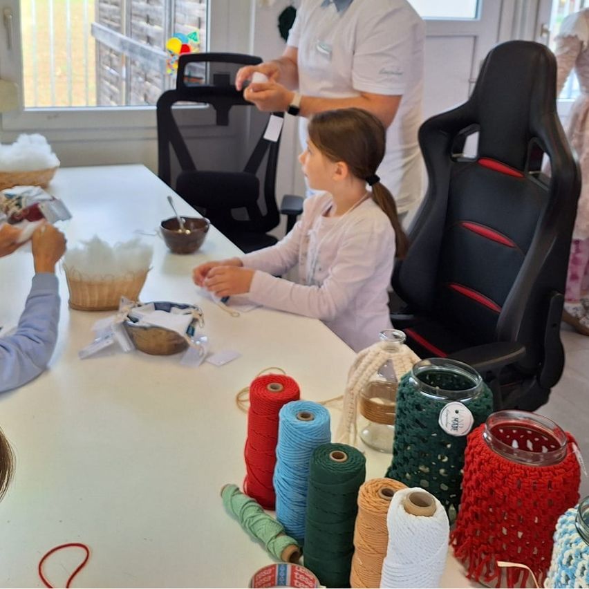 Ein Mann und ein kleines Mädchen arbeiten an einem Tisch, der mit Garn, Schüsseln und einem Korb gefüllt ist, während ein weiteres Kind zuschaut. Der Mann demonstriert der kleinen Mädchen etwas.