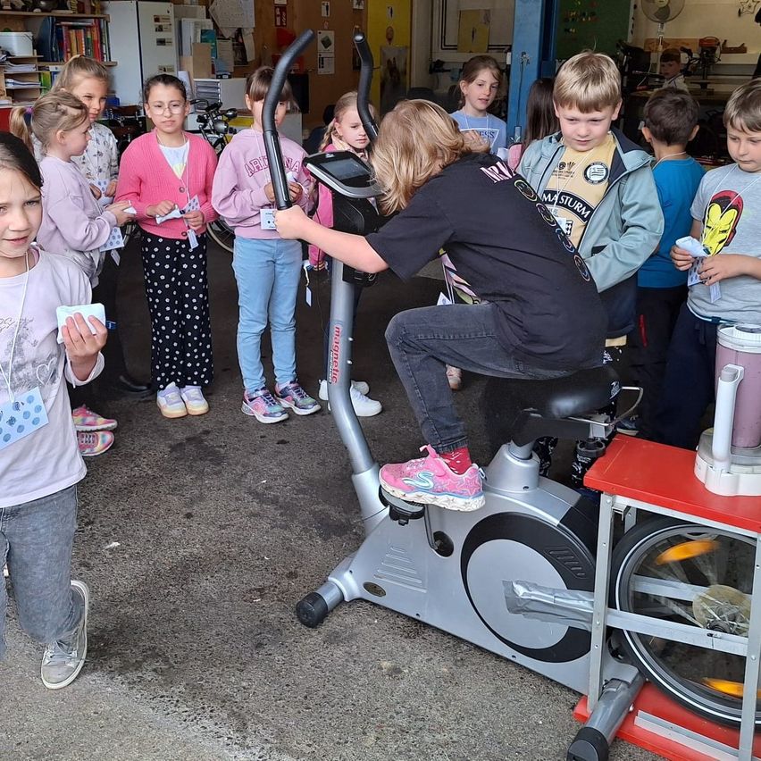 Eine Gruppe von Kindern steht um ein stationäres Fahrrad herum. Ein Kind tritt, während andere zuschauen. Einige halten Zettel in der Hand. Die Szene spielt sich in einem Raum mit Regalen und Fahrrädern im Hintergrund ab.