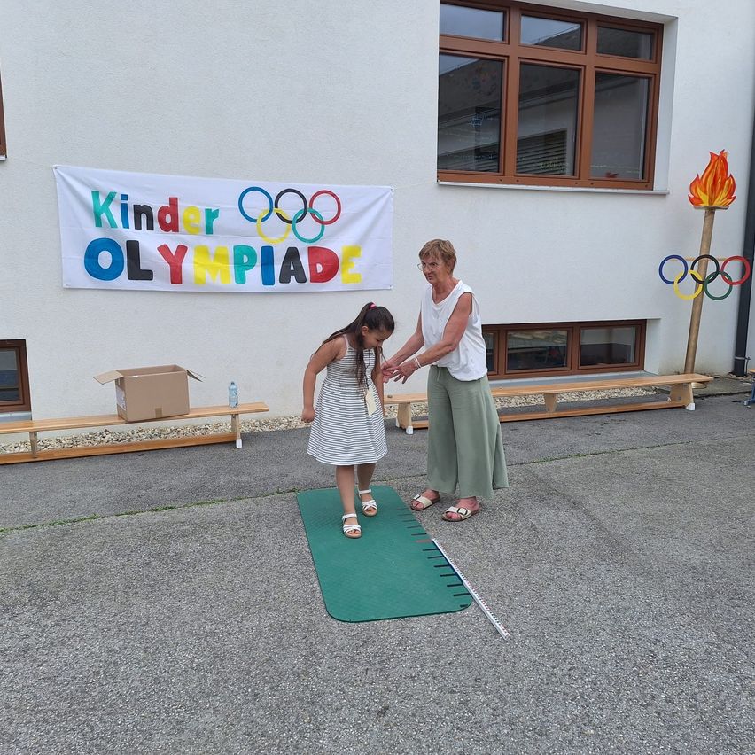 Eine erwachsene Frau führt ein junges Mädchen auf einem grünen Teppich vor einem Gebäude mit einem Banner, das 'Kinder Olympiade' steht. Ein Fackel steht in der Nähe.