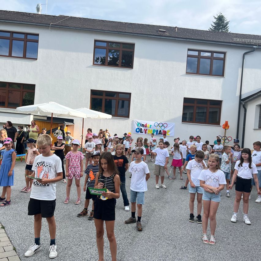Eine große Gruppe von Kindern in weißen T-Shirts steht in einem Kreis vor einem Gebäude mit einer olympischen Fahne. Die Kinder halten Medaillen und blicken in verschiedene Richtungen.