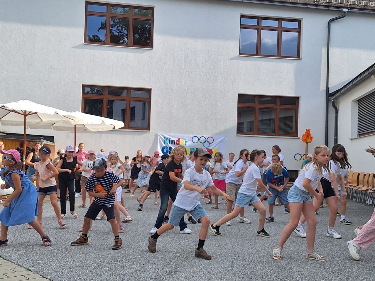 Kinder nehmen an einer Outdoor-Aktivität vor einem Gebäude mit weißer Wand und einem Banner, das die Kinder-Olympiade bewirbt, teil.