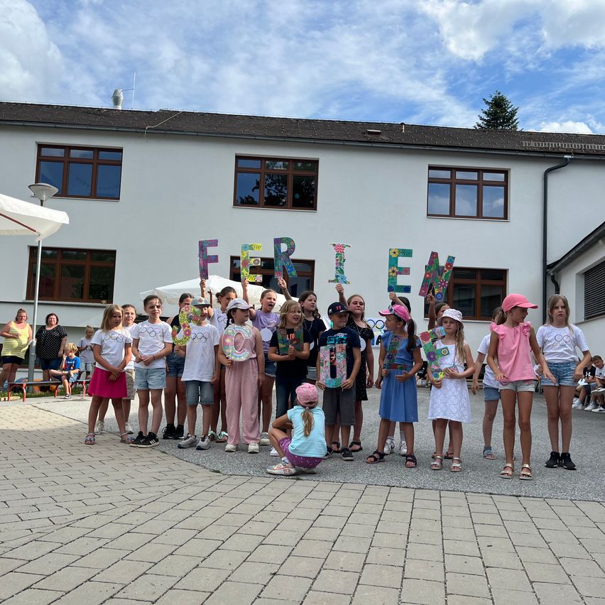 Eine Gruppe von Kindern steht vor einem Gebäude und hält Buchstaben, die 'FERIE' buchstabieren. Dahinter beobachten Erwachsene. Das Gebäude hat viele Fenster und einen klaren Himmel darüber.