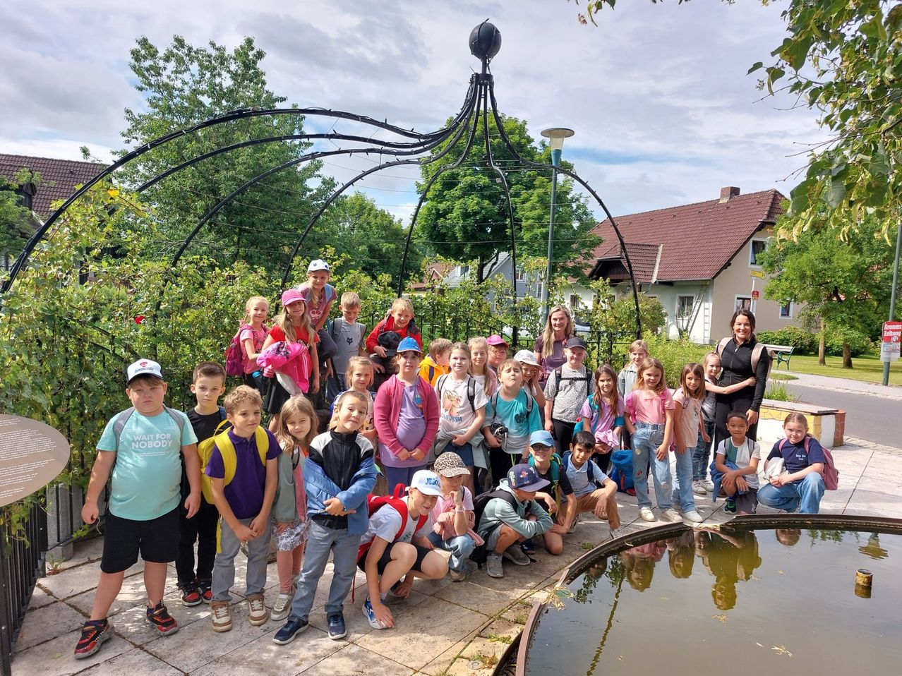 Eine Gruppe von Kindern und Erwachsenen versammelt sich draußen in einer Gartenumgebung, einige stehen, einige sitzen und eine Frau steht hinter ihnen. Sie scheinen auf einem Ausflug zu sein.