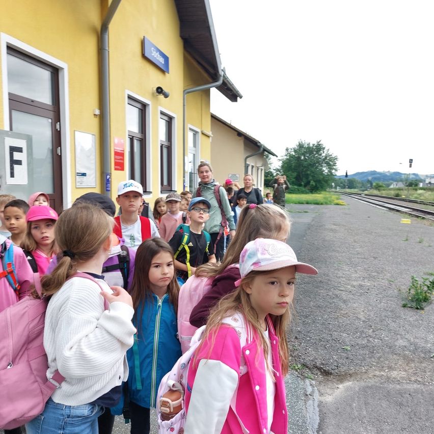 Eine Gruppe von Kindern und ihren Begleitpersonen steht an einem Bahnhofsgebäude und blickt auf die Gleise. Einige Kinder tragen Hüte und haben Rucksäcke.