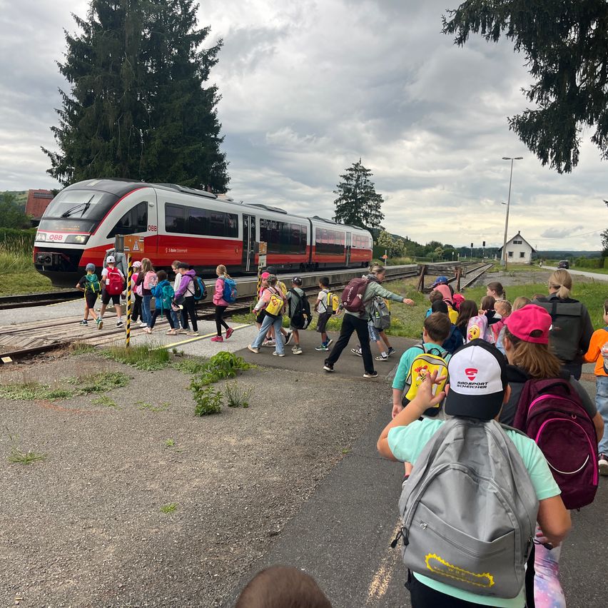 Eine Gruppe von Schulkindern in Uniformen und Rucksäcken überquert eine Bahnlinie, während ein Zug herankommt. Der Zug ist rot und weiß, und im Hintergrund befinden sich Bäume und ein kleines Haus.