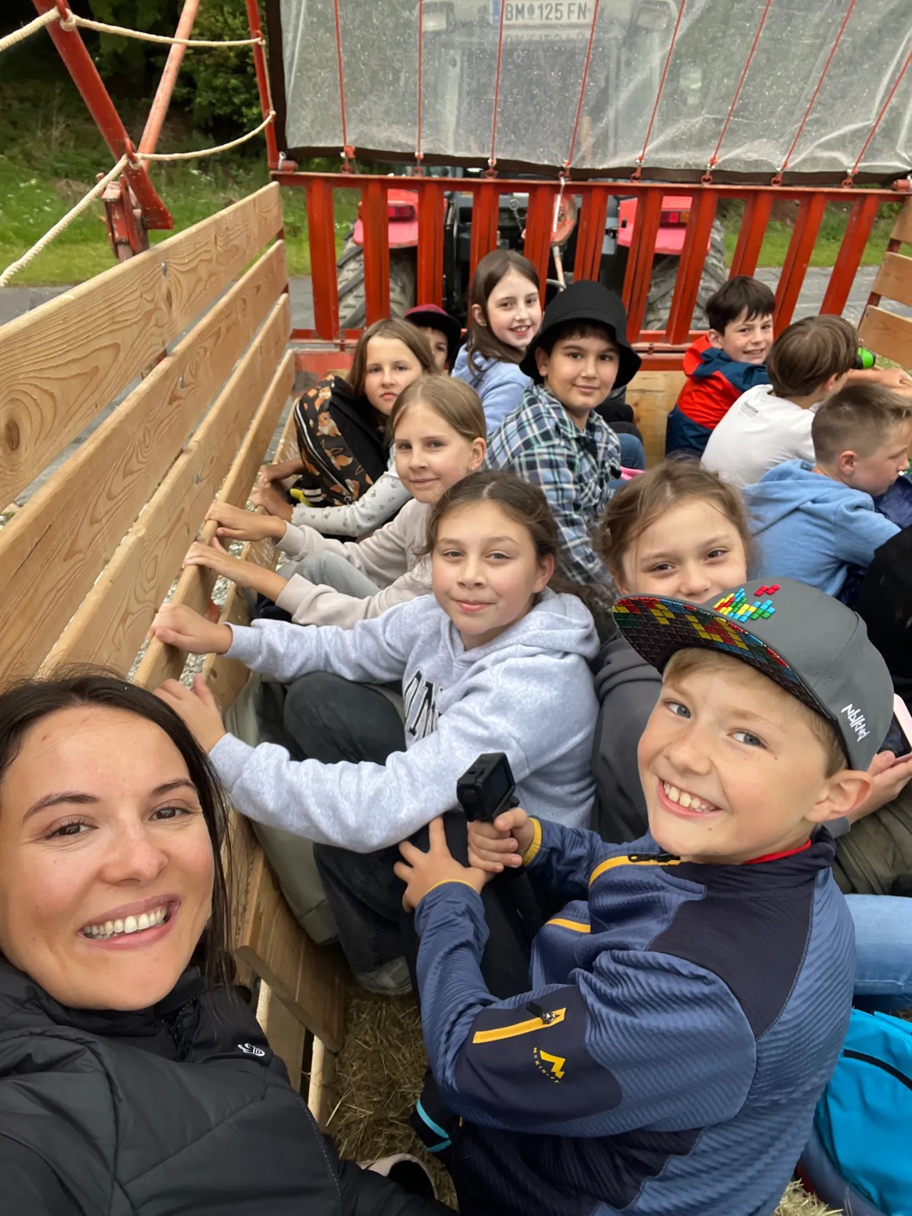 Eine Gruppe von Kindern sitzt in einem Traktoranhänger mit einer lächelnden erwachsenen Frau. Die Kinder tragen Freizeitkleidung und einige tragen Hüte. Der Anhänger ist aus Holz und sie befinden sich auf einem Bauernhof.