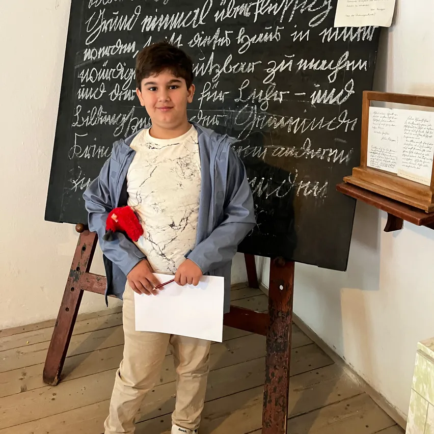 Ein junger Junge hält ein Papier und einen Stift in der Hand und steht vor einer Tafel mit kursiver Schrift und einem kleinen gerahmten Dokument rechts von ihm.