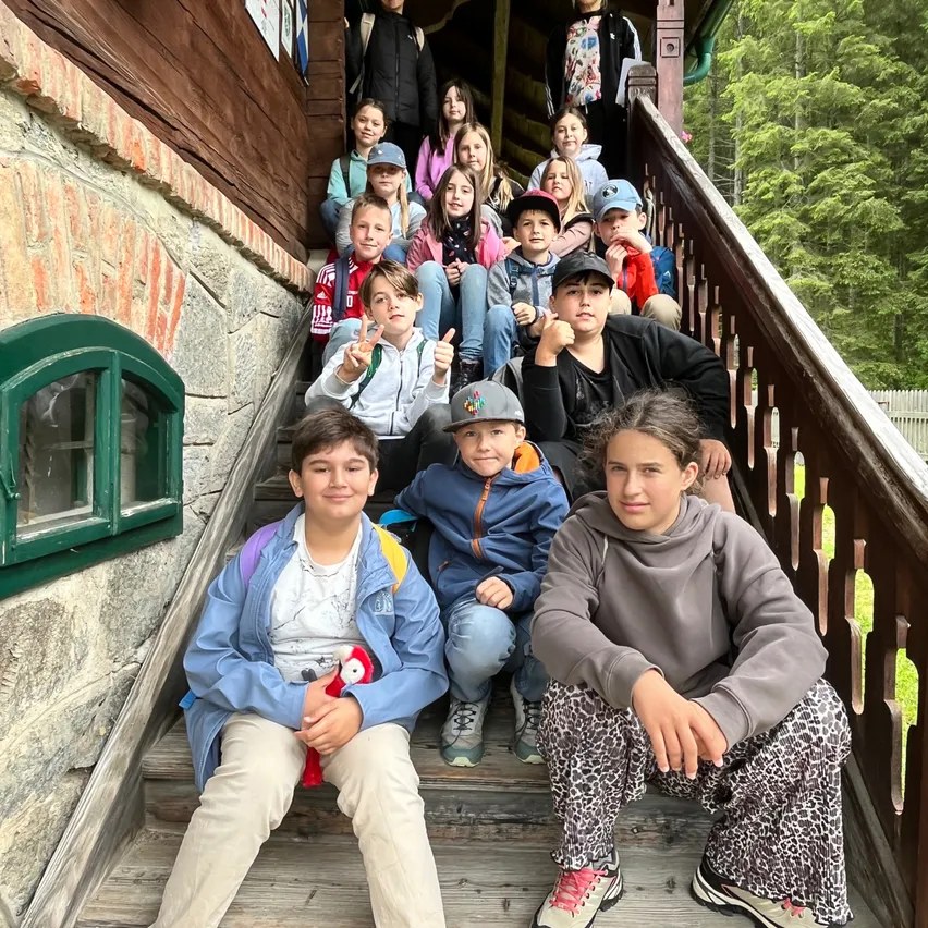 Eine Gruppe von Kindern sitzt auf den Stufen eines Holzhauses mit einer Ziegelmauer und einem grünen Fenster. Sie lächeln für ein Foto.