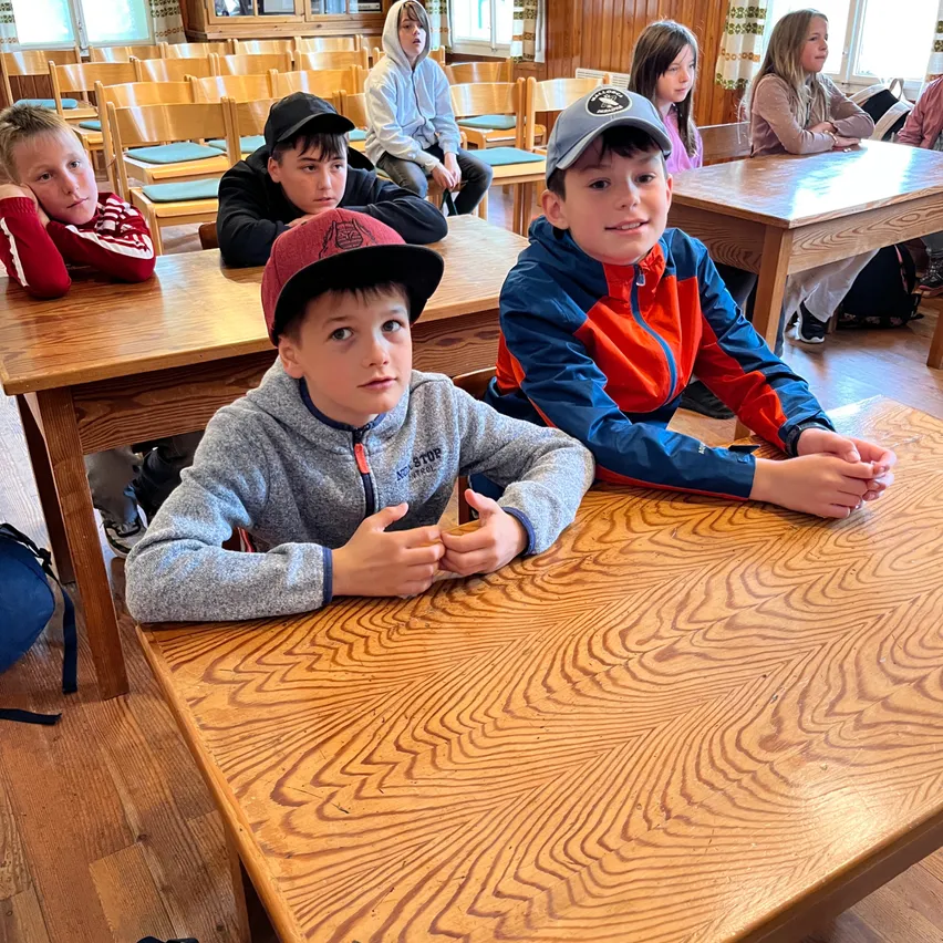 Fünf Jungen sitzen an Holztischen in einem Klassenzimmer, einer in einem roten Shirt, ein anderer in einem grauen Hoodie, ein Junge in einem blauen Jackett und zwei weitere in Hüten.