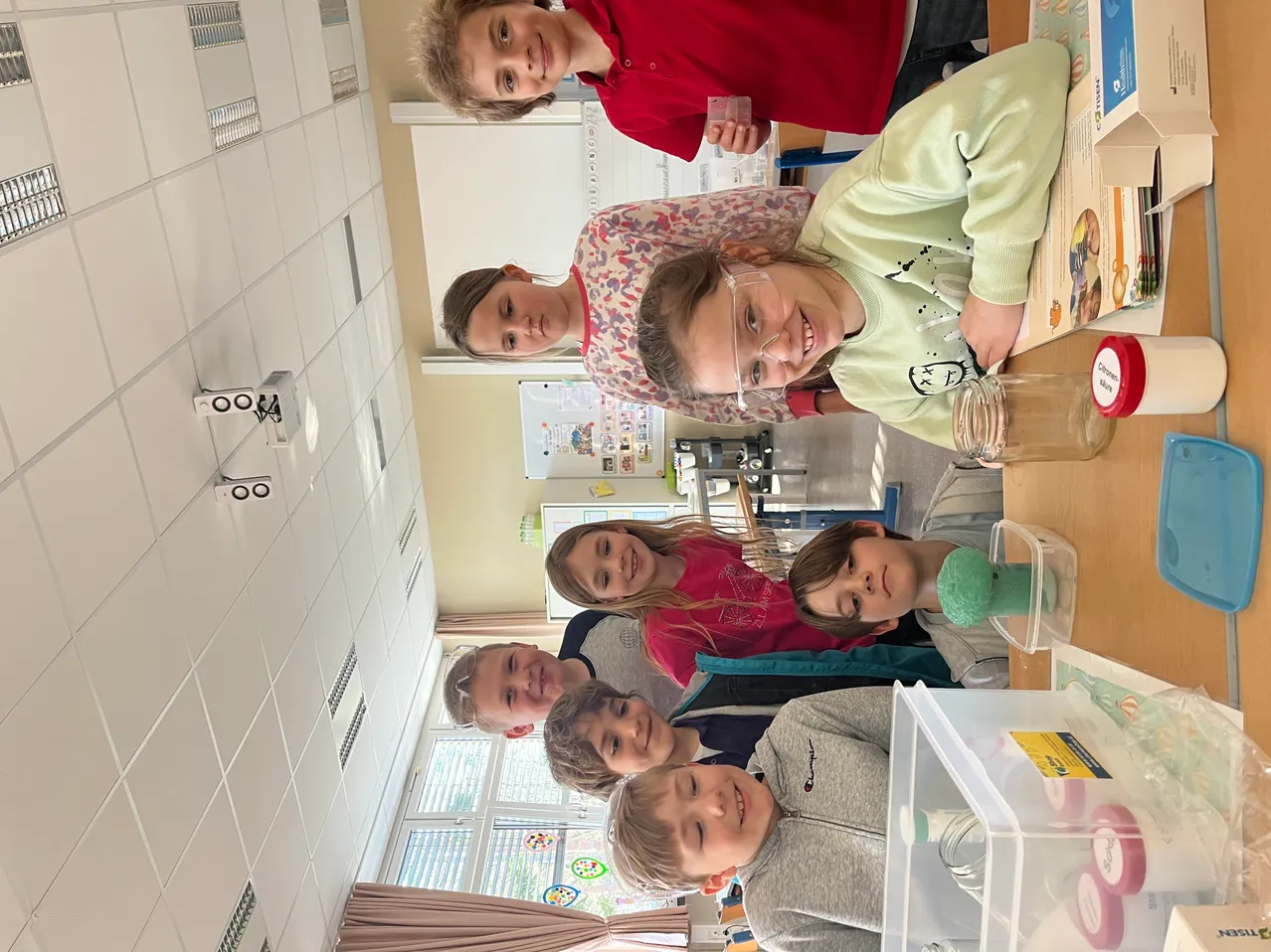 Eine Gruppe von Kindern in einem Klassenzimmer mit einem Glas auf einem Schreibtisch, lächeln und schauen zur Kamera. Ein Kind hält ein grünes Objekt.