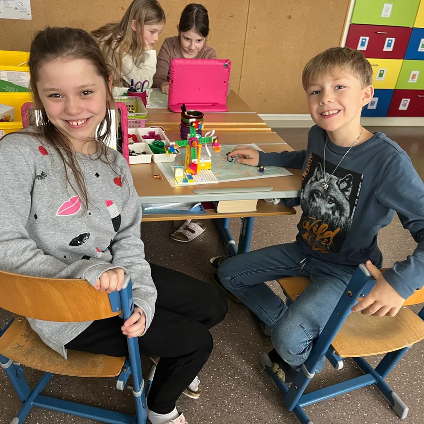 Zwei Kinder sitzen an einem Tisch, eines baut mit Lego. Hinter ihnen sitzen weitere Kinder und arbeiten mit Laptops und anderen Materialien.