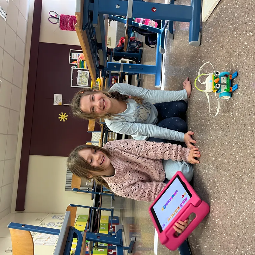Zwei junge Mädchen sitzen im Klassenzimmer auf dem Boden, lächeln und posieren für ein Foto. Eine von ihnen hält ein Tablet. In der Nähe steht ein Spielzeugroboter auf dem Boden.